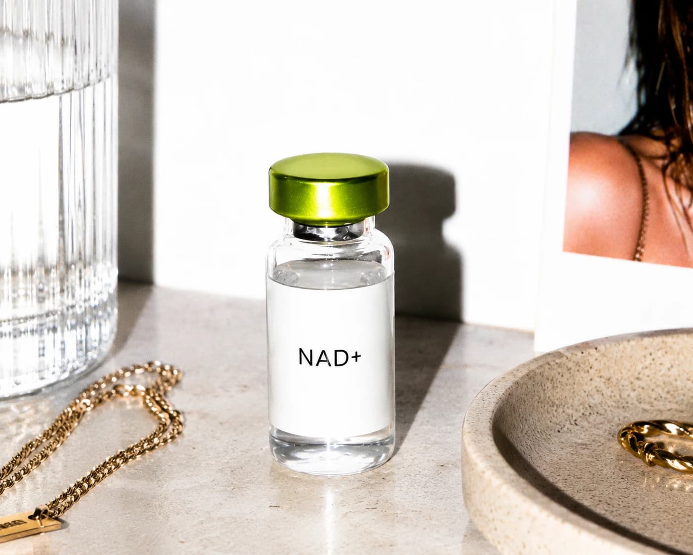 An Atelier Protocol NAD+ vial with a green cap on a sunlit stone surface beside gold chain jewelry, a ribbed glass of water, and a framed photo