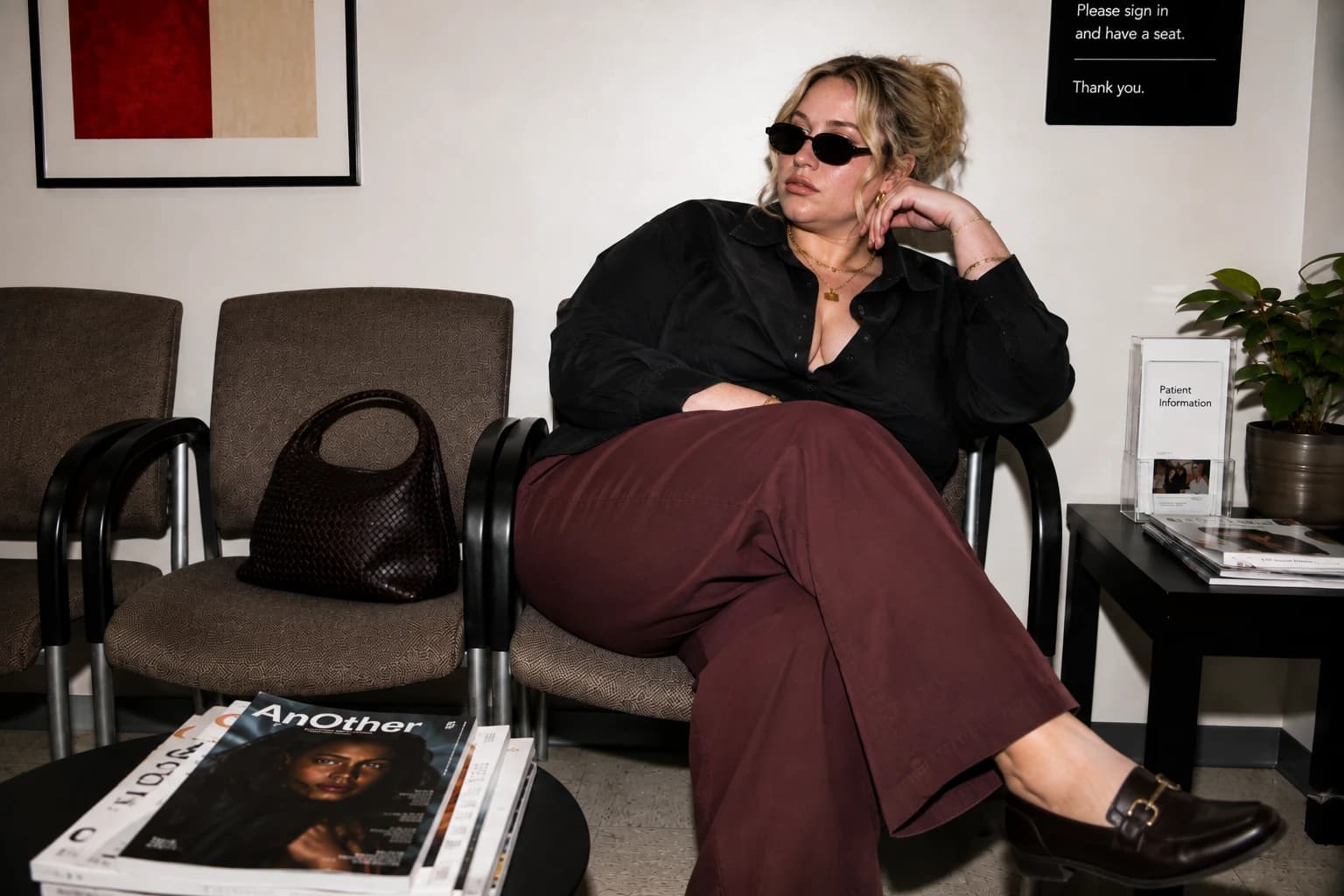 A woman in slim sunglasses, a black satin shirt, and burgundy trousers relaxing in a calm, considered waiting space