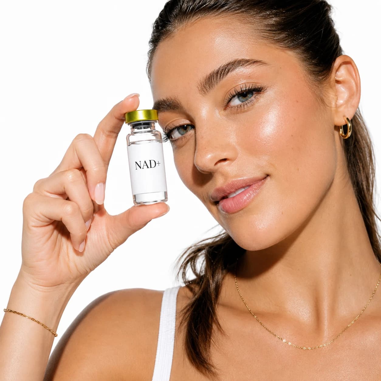 A woman with glowing skin and gold jewelry holding a gold-cap NAD+ vial beside her eye on a bright white background.